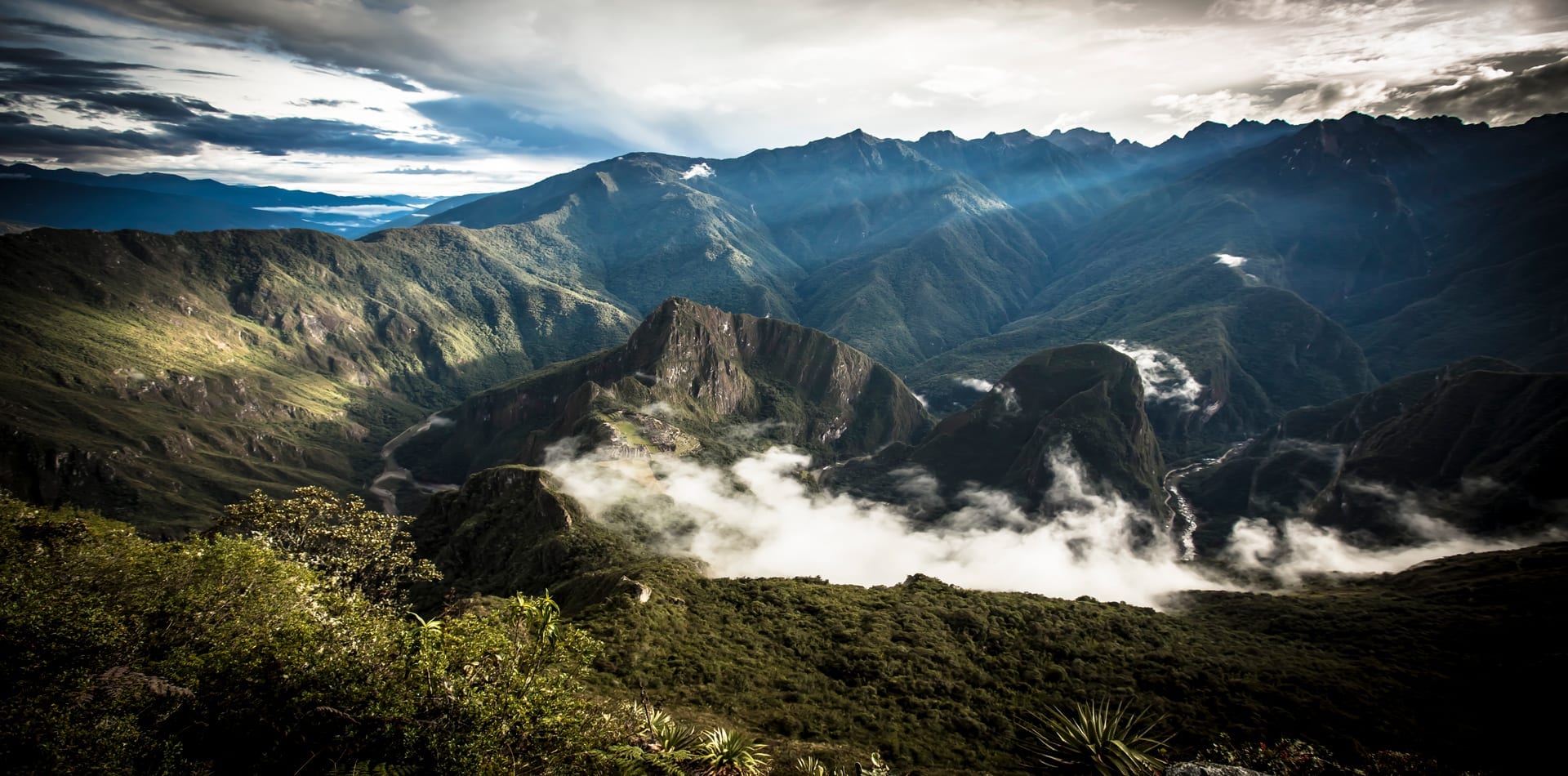 Panoramic view of the Machu Picchu ruins nestled between steep, green mountains and wispy clouds.