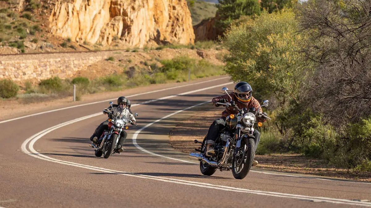 Two motorcyclists leaning into a curve on a sunlit road bordered by dry brush and rocky orange cliffs.