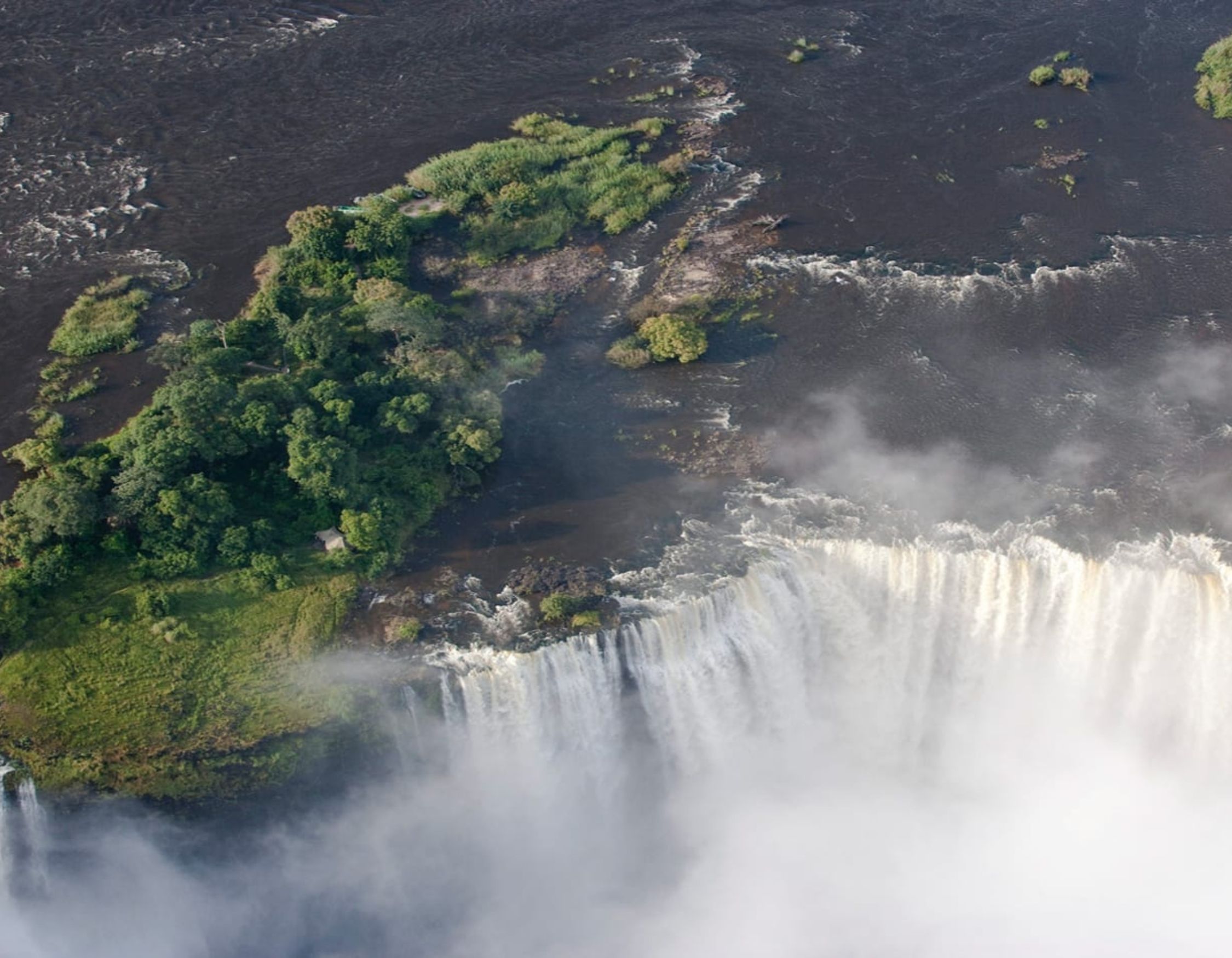 Aerial view of a large waterfall plunging into a mist-filled canyon with a lush, green island at the edge.