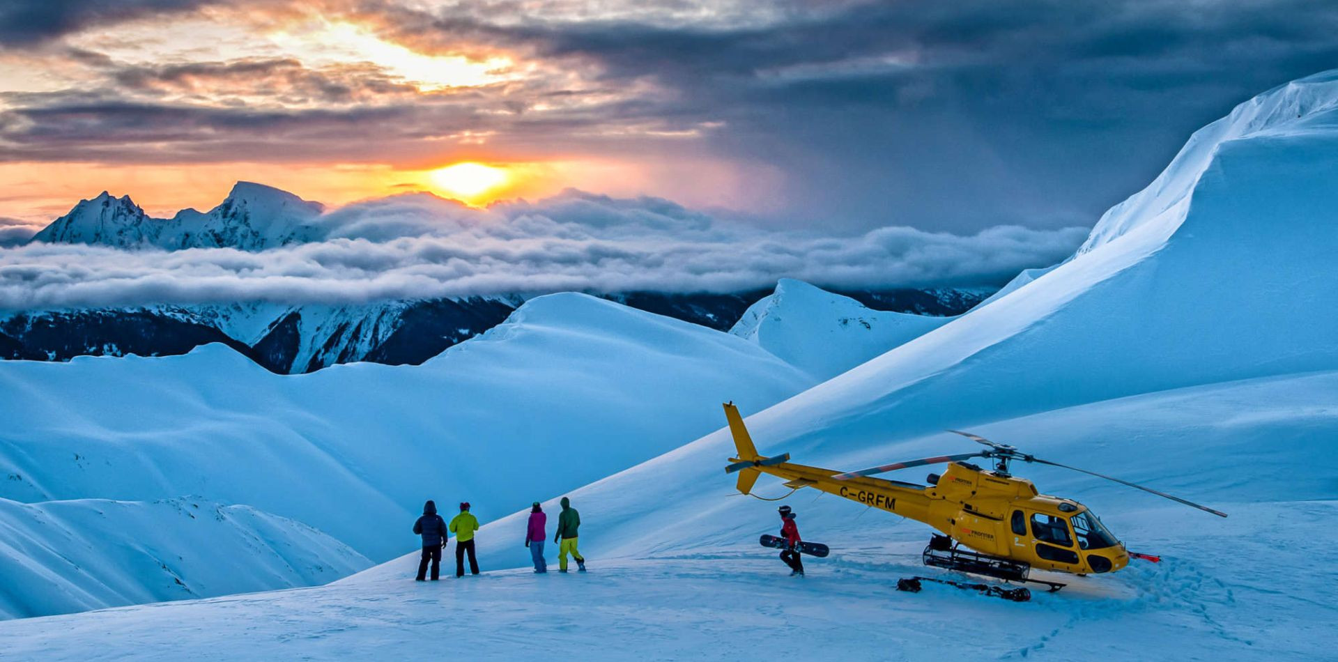 Skier walking toward a yellow helicopter on a snowy mountain ridge during a vibrant orange sunset.