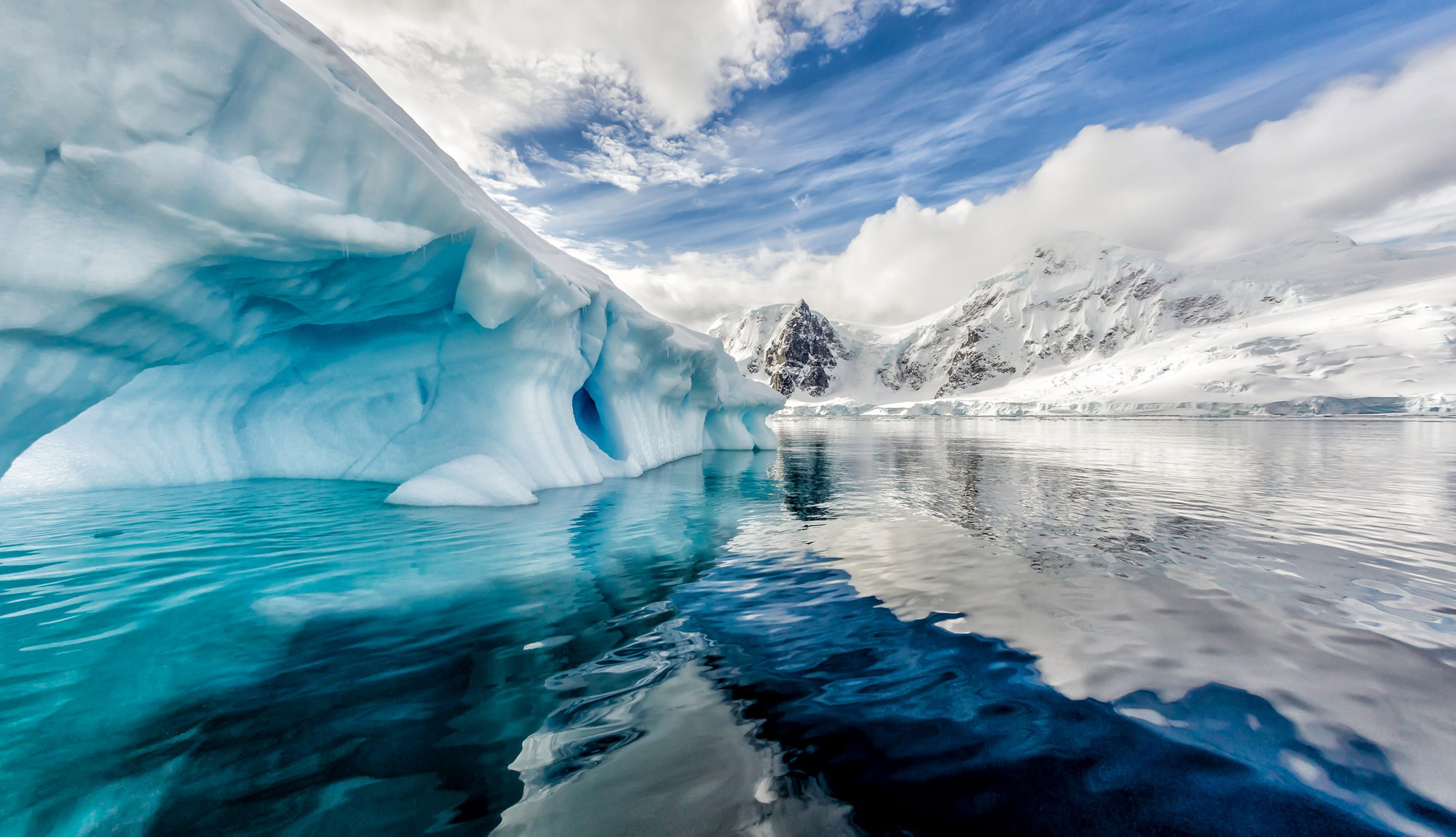 Massive blue-tinted icebergs floating in calm, reflective water under a bright sky with wispy clouds.