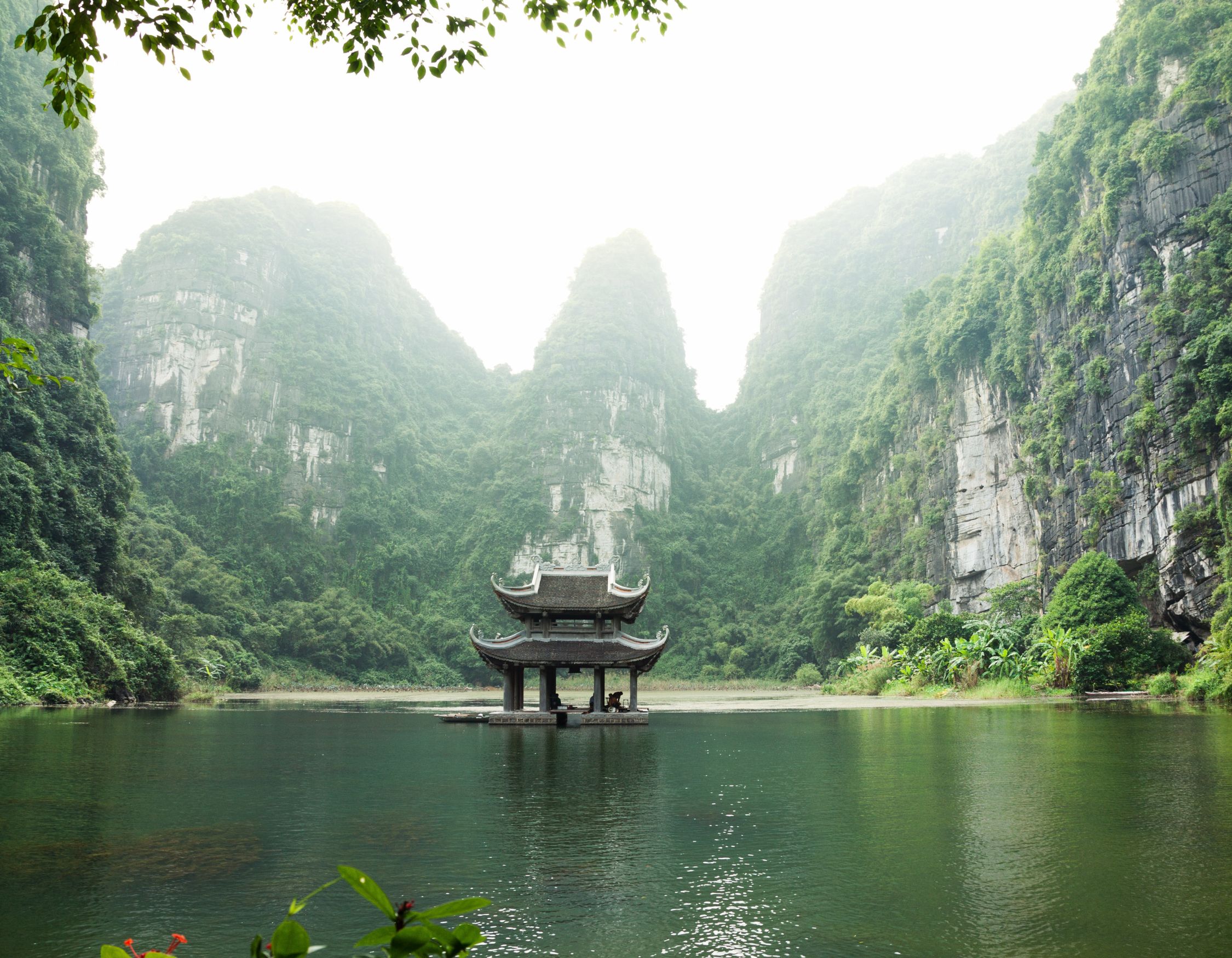 Ancient stone temple with a multi-tiered roof situated on a calm lake surrounded by limestone karst mountains.