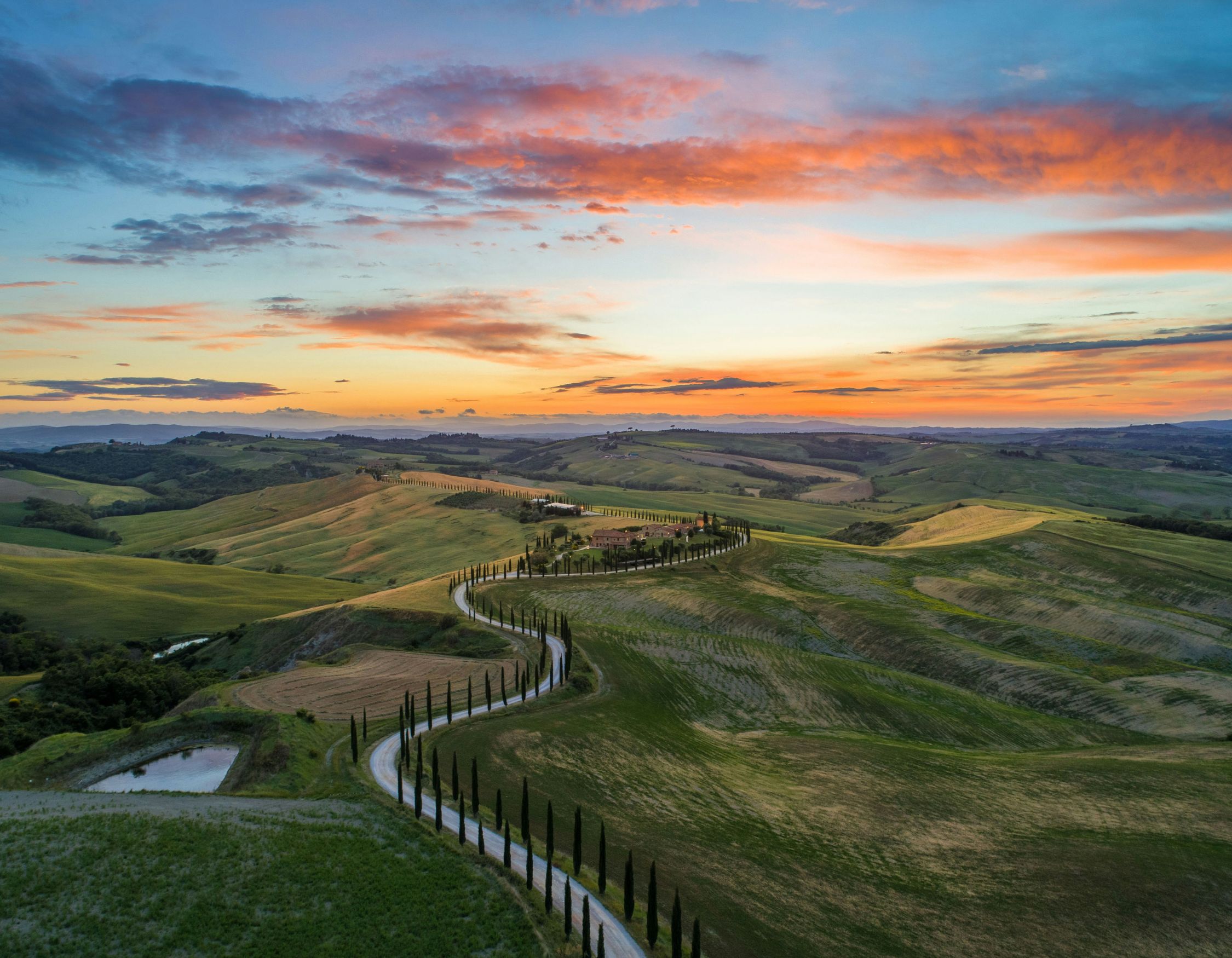 Aerial view of a winding road lined with cypress trees leading to a villa in the rolling hills of Tuscany at sunset.
