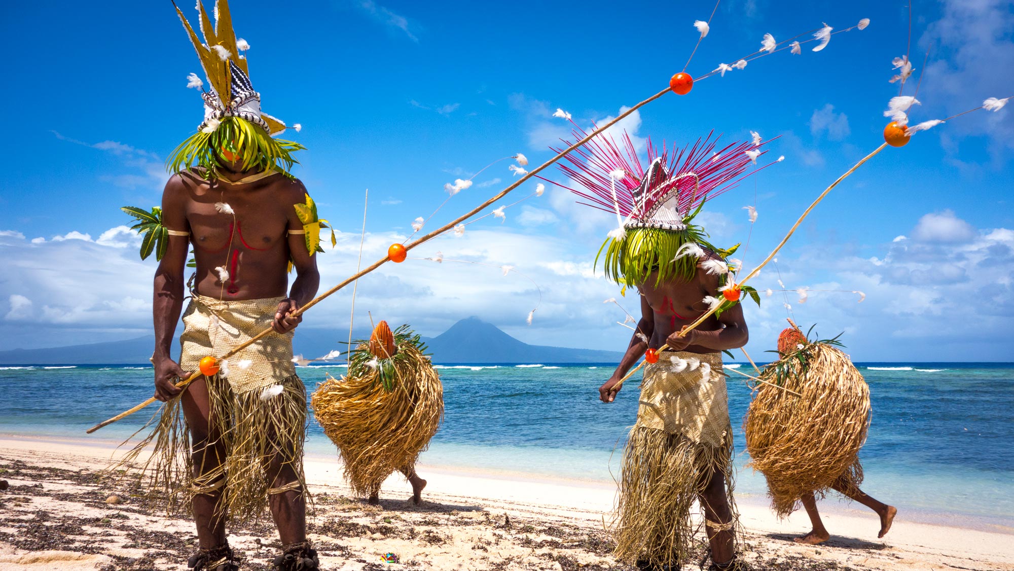 Men in traditional indigenous attire with elaborate feathered headpieces dancing on a white sand beach by the ocean.