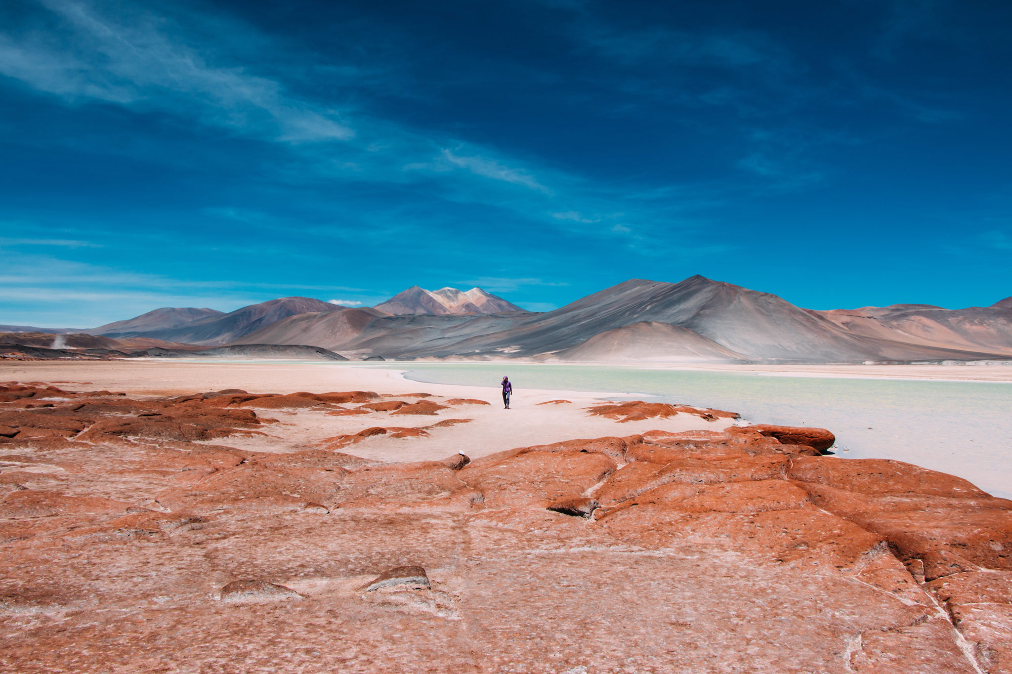 Lone hiker walking across a white salt flat toward distant mountains under a bright blue sky.