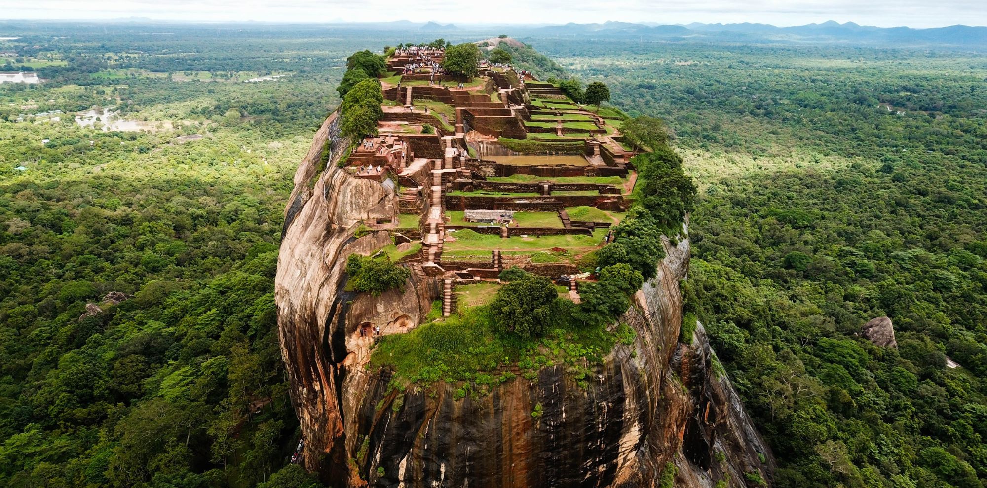 Aerial view of Sigiriya Rock Fortress in Sri Lanka, showing terraced ruins atop a massive granite column surrounded by jungle.