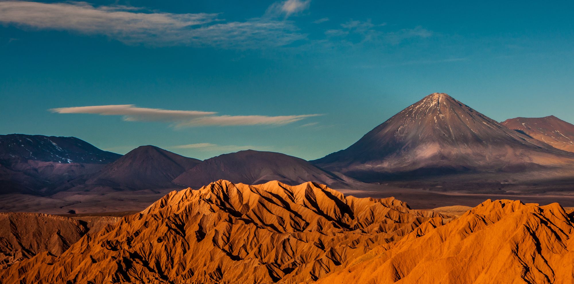 Jagged orange rock formations in the foreground with a towering, perfectly shaped volcanic peak in the distance.