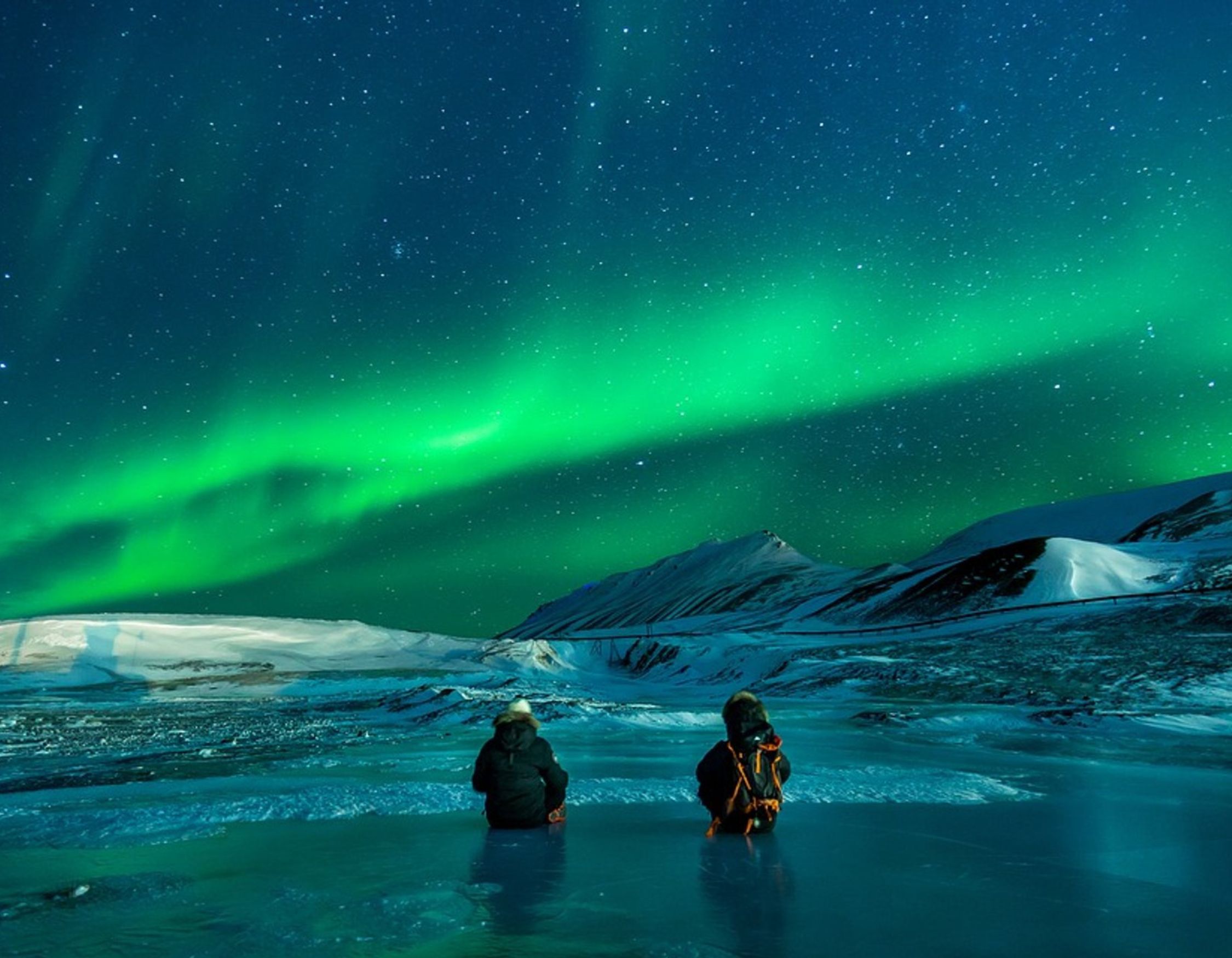Two people sitting on a frozen lake watching a vibrant green aurora borealis stretch across a starry night sky.