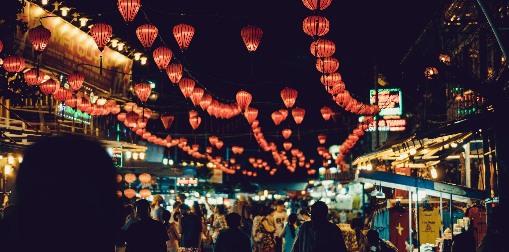 Rows of glowing red paper lanterns hanging over a crowded night market street filled with people and stalls.