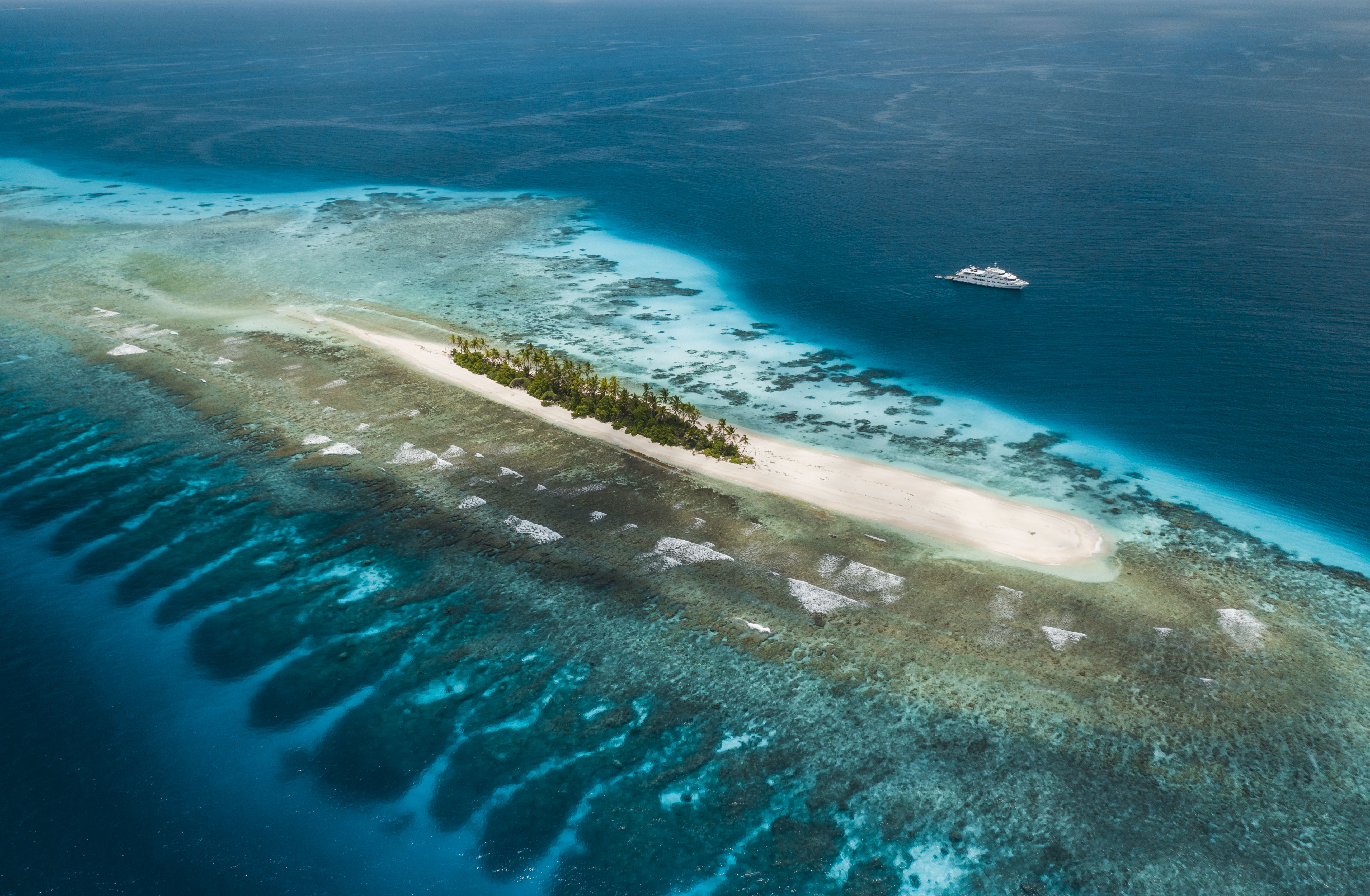 An aerial view of a small, sandy island surrounded by vibrant turquoise waters and coral reefs. Lush green palm trees dot the island, while a yacht floats in the distance.