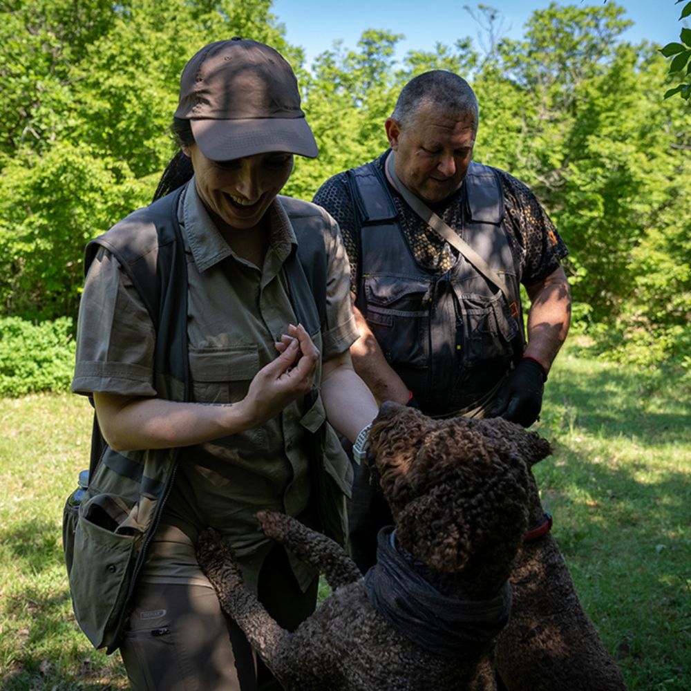 A woman in a khaki outfit interacts with a dog while a man in a dark vest watches, both smiling in a green forested area.