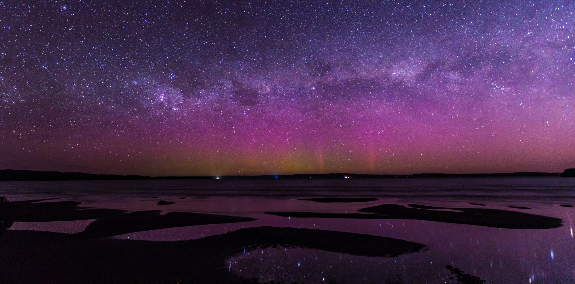 A stunning night sky filled with a vibrant display of stars and the Milky Way, set above a calm body of water. The sky features hues of purple, pink, and green, reflecting subtly on the water's surface. Silhouettes of land and shapes are visible in the foreground.
