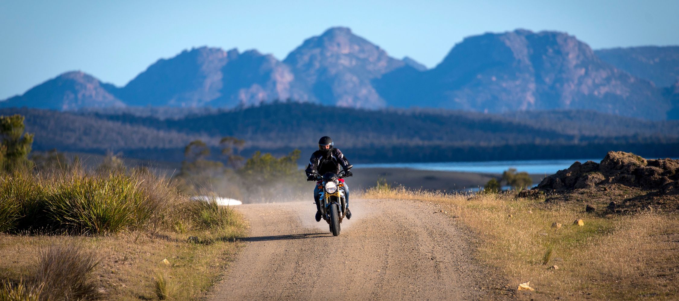 A motorcyclist rides along a dirt road with mountains in the background and a body of water visible nearby. Bright sunlight illuminates the scene, creating a sense of adventure in a picturesque landscape.