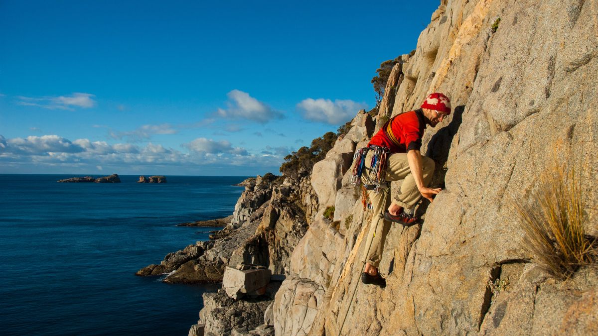 A climber ascends a rugged rock face against a backdrop of a clear blue sky and calm ocean. The coastline features rocky outcrops in the distance.