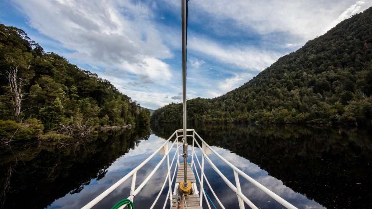 A tranquil scene of a boat's deck extending into a calm river, flanked by dense green hills under a cloudy sky. The water reflects the surrounding nature, creating a symmetrical view.