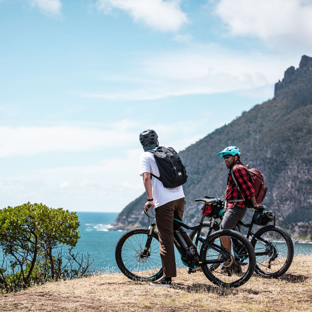 Two mountain bikers stand on a grassy overlook, gazing at a scenic ocean view. One wears a light shirt and black backpack, while the other sports a plaid shirt and a blue helmet. A coastline and mountains are visible in the background.