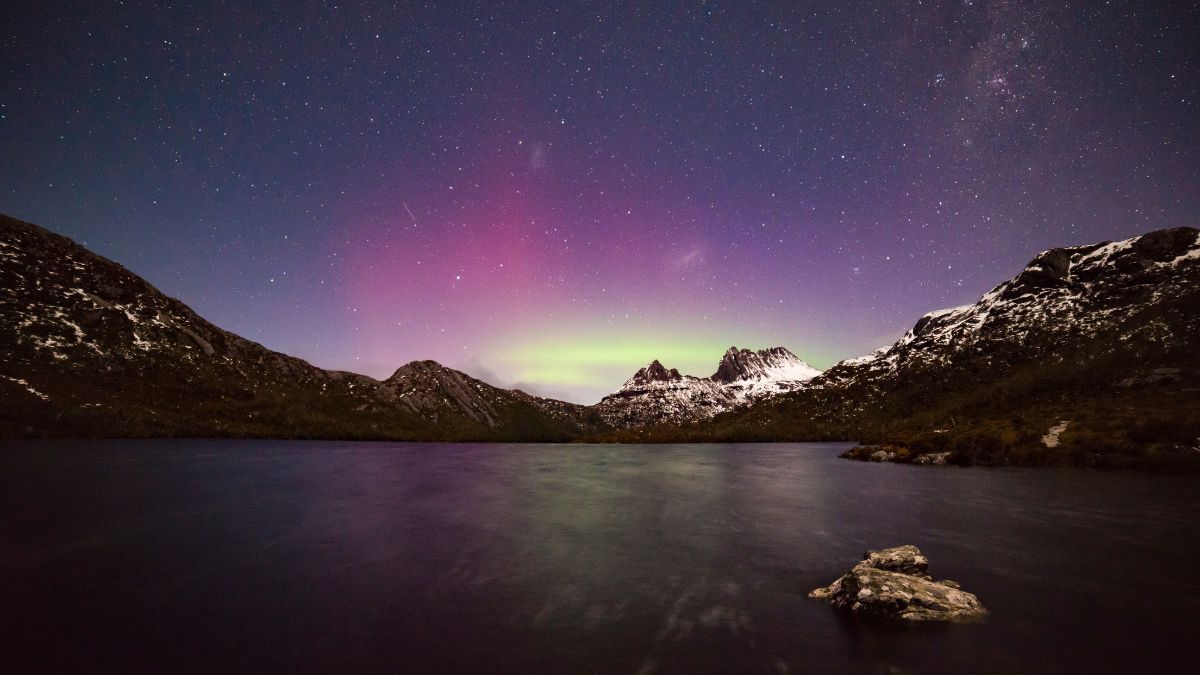 A tranquil lake reflects the night sky illuminated by vibrant auroras, with rugged mountains in the background and stars twinkling above.