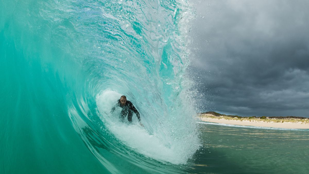A surfer rides a large, turquoise wave, partially submerged in the water with a cloudy sky in the background. The scene captures a dynamic moment of surfing, showcasing the power of the ocean.