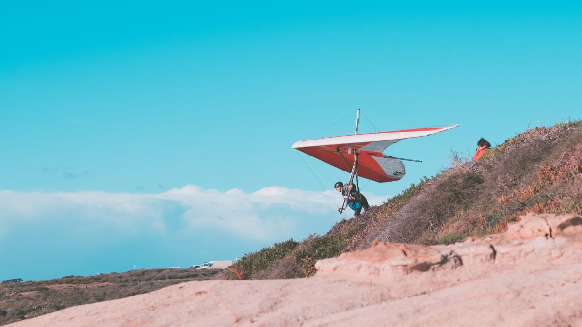 A person carrying a hang glider prepares to take off from a rocky hillside against a clear blue sky. Another person stands nearby, watching the preparation.