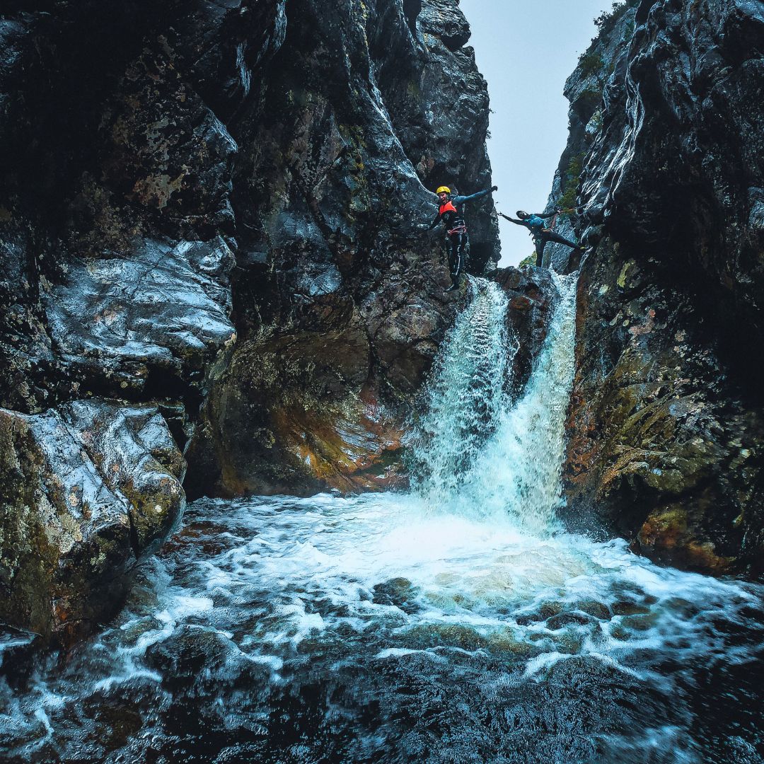 Two climbers navigating a narrow canyon with rushing water below. One climber is descending a cliff while the other assists from a ledge. Mist and rugged rock formations surround the scene.