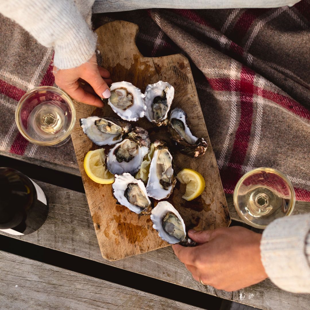 Hands reaching for fresh oysters on a wooden platter, garnished with lemon slices, accompanied by two glasses of white wine on a rustic table. A cozy blanket can be seen in the background.