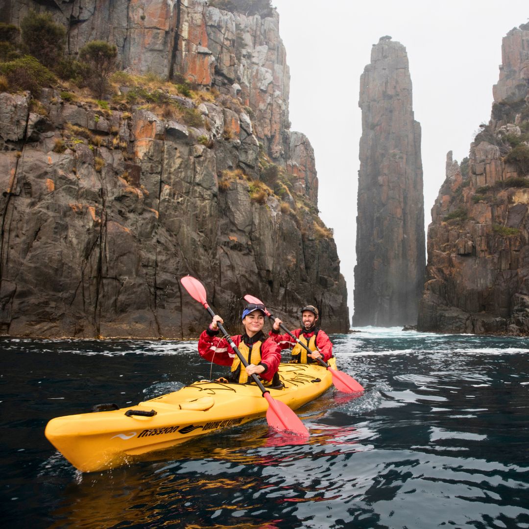 Two kayakers paddle through a narrow waterway between towering rock formations. The rocky cliffs are covered in greenery, and a mist shrouds the top of the cliffs. The kayakers wear yellow life vests and are smiling, enjoying their adventure.