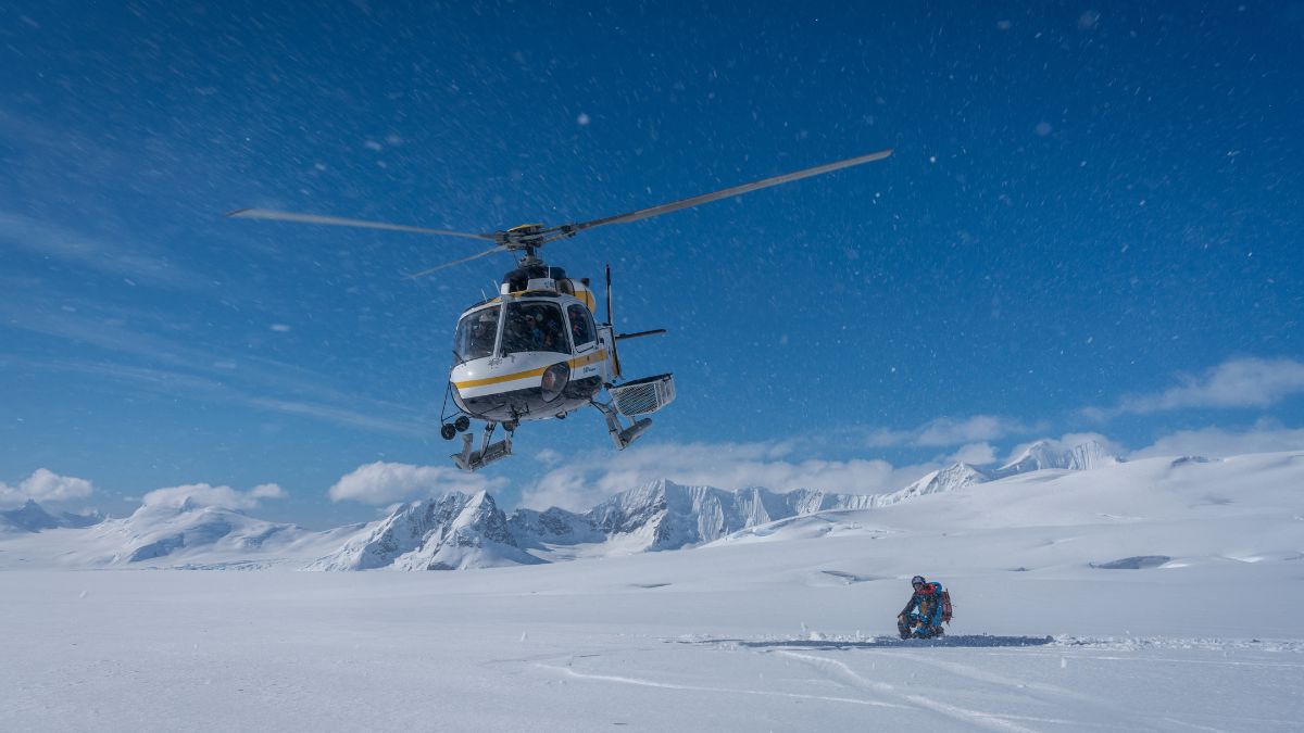 A white and yellow helicopter lifting off from a flat, snow-covered landscape with jagged mountains in the distance.