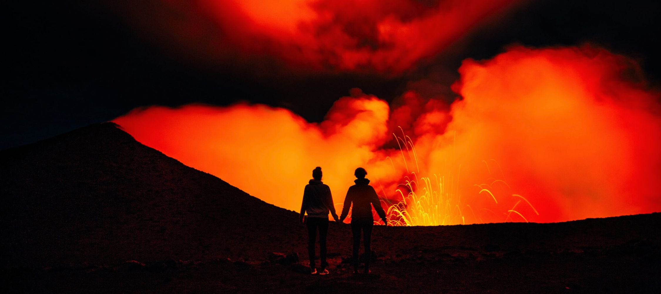 A silhouetted couple holding hands in front of an erupting volcano, surrounded by bright orange lava and thick, glowing smoke against a dark sky.