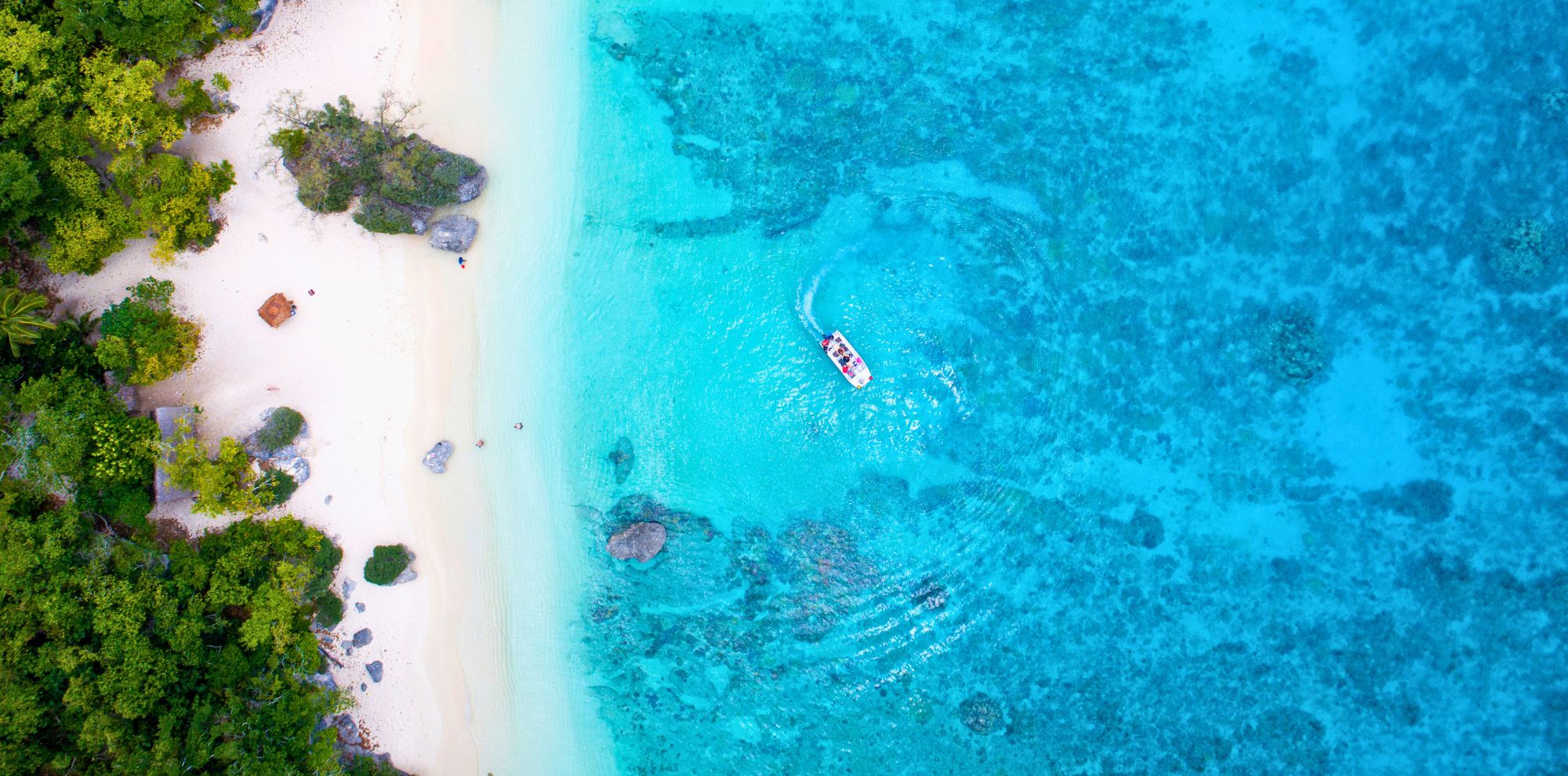Aerial view of a tropical beach with white sand and vibrant turquoise water, showing a boat moving through the clear water near the shore. Lush green vegetation lines the beach.
