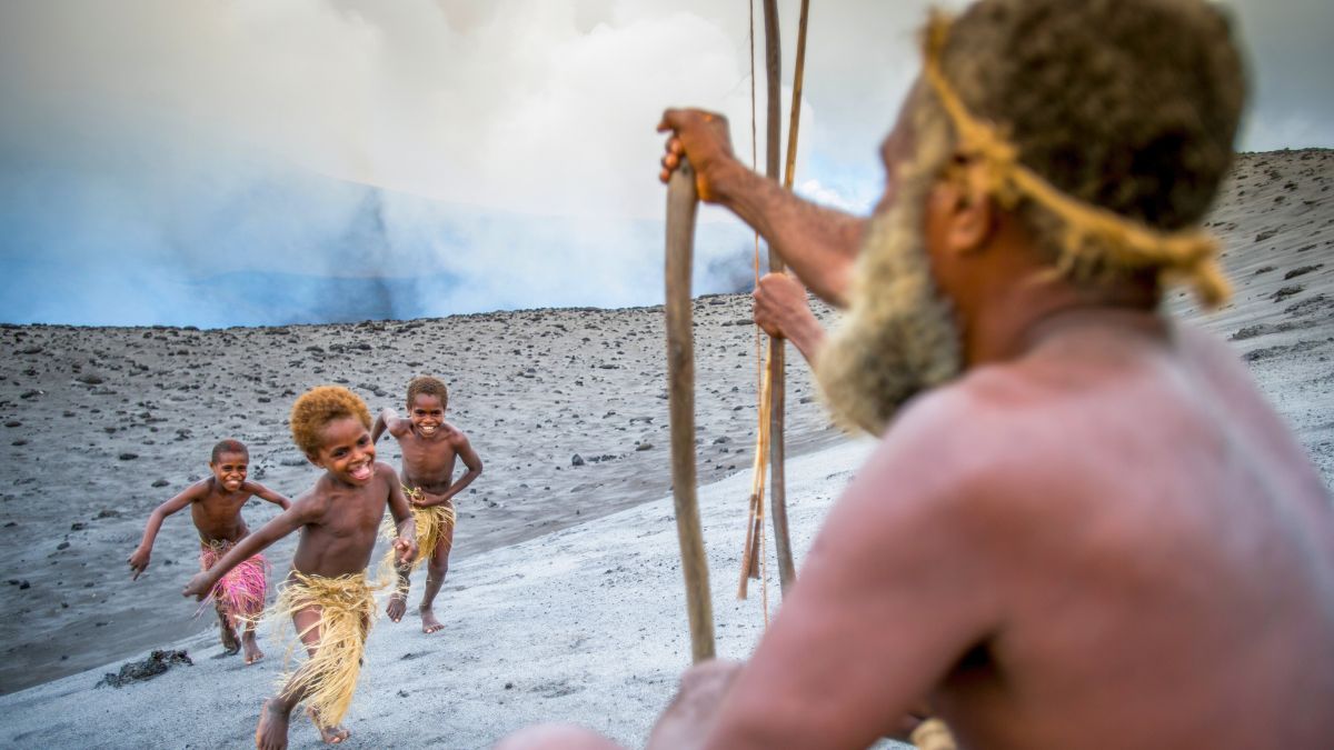 A group of four children playfully run toward an elderly man sitting on a volcanic landscape. The children have light brown skin and wear traditional grass skirts, while the man has a long beard and is holding a spear. Smoke rises from the volcanic background, enhancing the adventurous atmosphere.