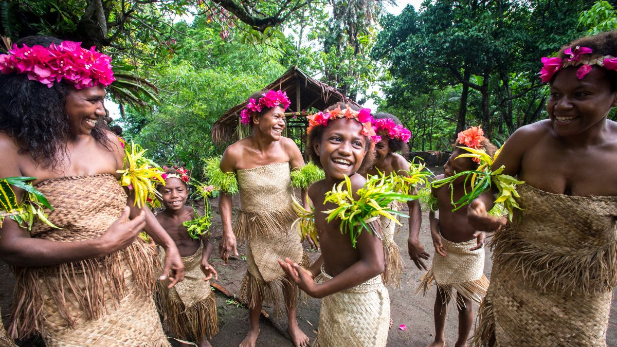 Women and children in traditional attire are joyfully dancing outdoors, adorned with vibrant flower crowns and holding greenery. Behind them, a rustic thatched hut and lush foliage create a natural backdrop.