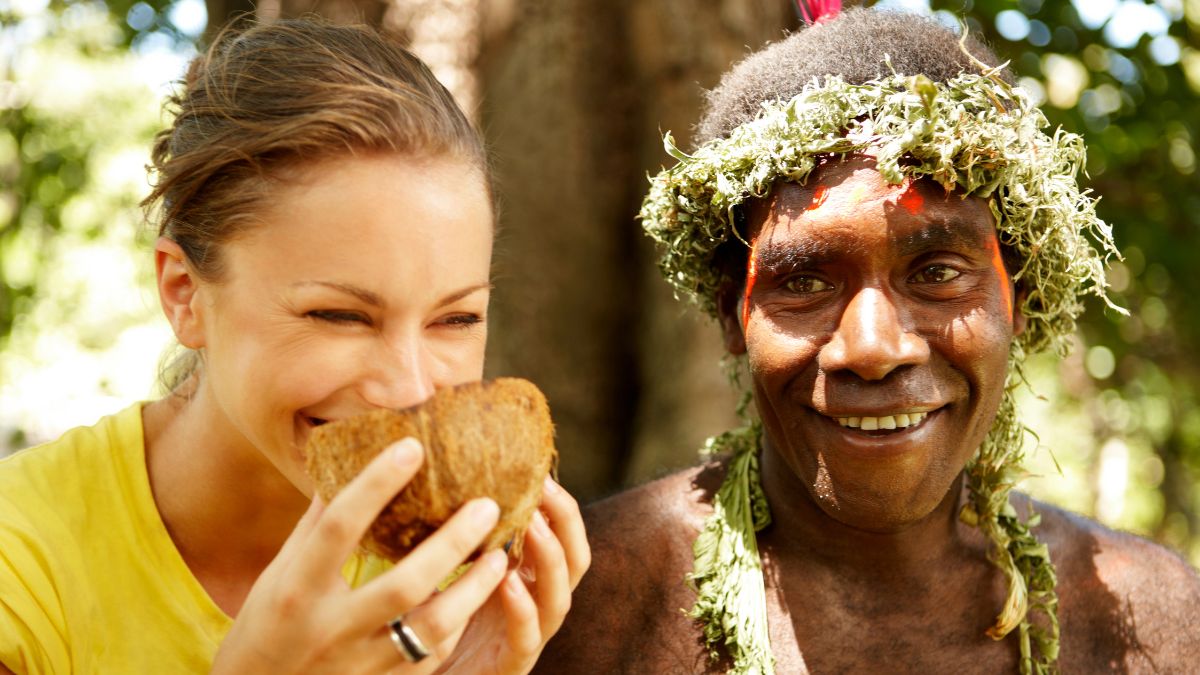 A woman holding a traditional food item smiles at a man adorned with foliage and a headdress, both enjoying a moment together in a lush outdoor setting.