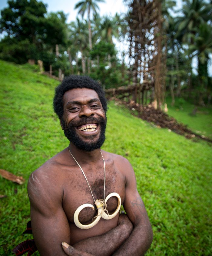 A smiling man with a beard and long straight hair stands with crossed arms, wearing a necklace made of wooden rings. He is surrounded by lush greenery, with trees in the background.