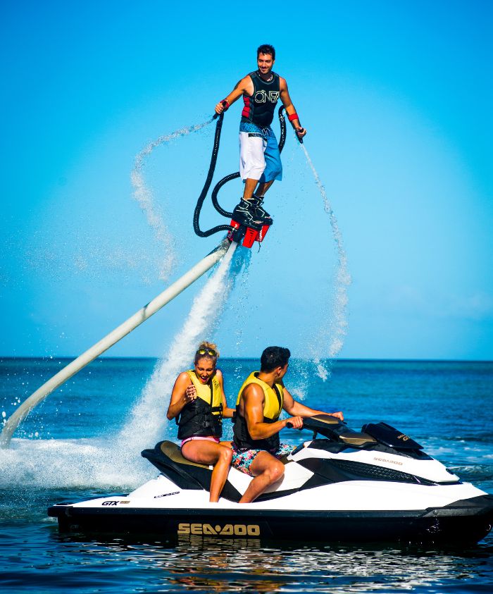 A person using a flyboard shoots up into the air above a body of water, while two people ride a jet ski nearby, enjoying the sunny day.