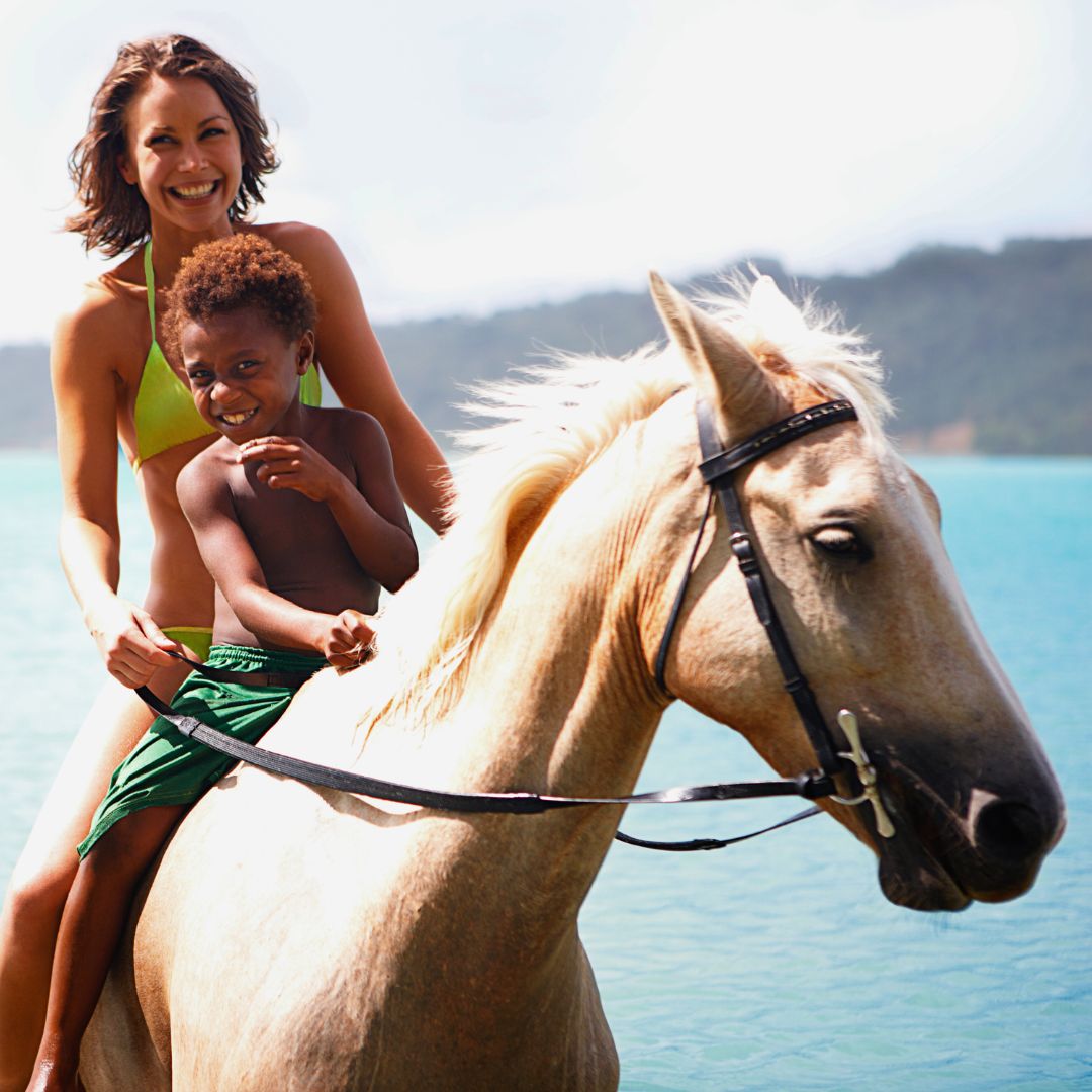 A woman in a bikini smiles while riding a light-colored horse, with a young boy joyfully sitting in front of her. They are by a calm body of water, surrounded by greenery.