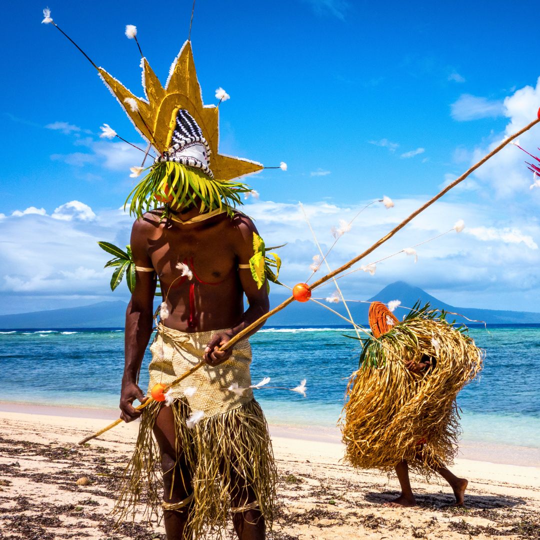 Two individuals in traditional attire standing on a beach. One wears a large, decorative headdress and holds a spear, while the other carries a bundle of grass. The background features a vibrant blue sky and water, with a distant mountain visible.