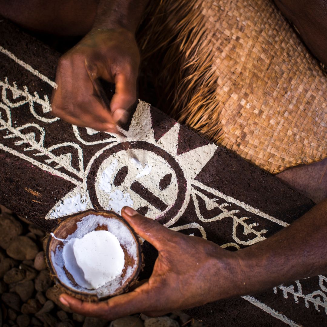 A person's hands are seen applying white powder to a traditional patterned surface, while a small bowl of white powder is nearby. The intricate design features a sun motif.