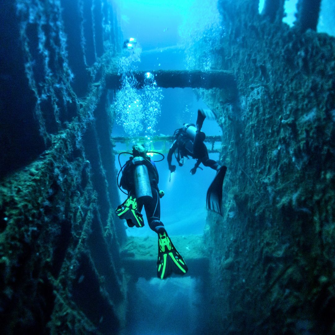 Two divers exploring an underwater wreck, surrounded by bubbles and the blue hues of the ocean. The wreck shows structures covered in marine growth, creating a sense of adventure and discovery.