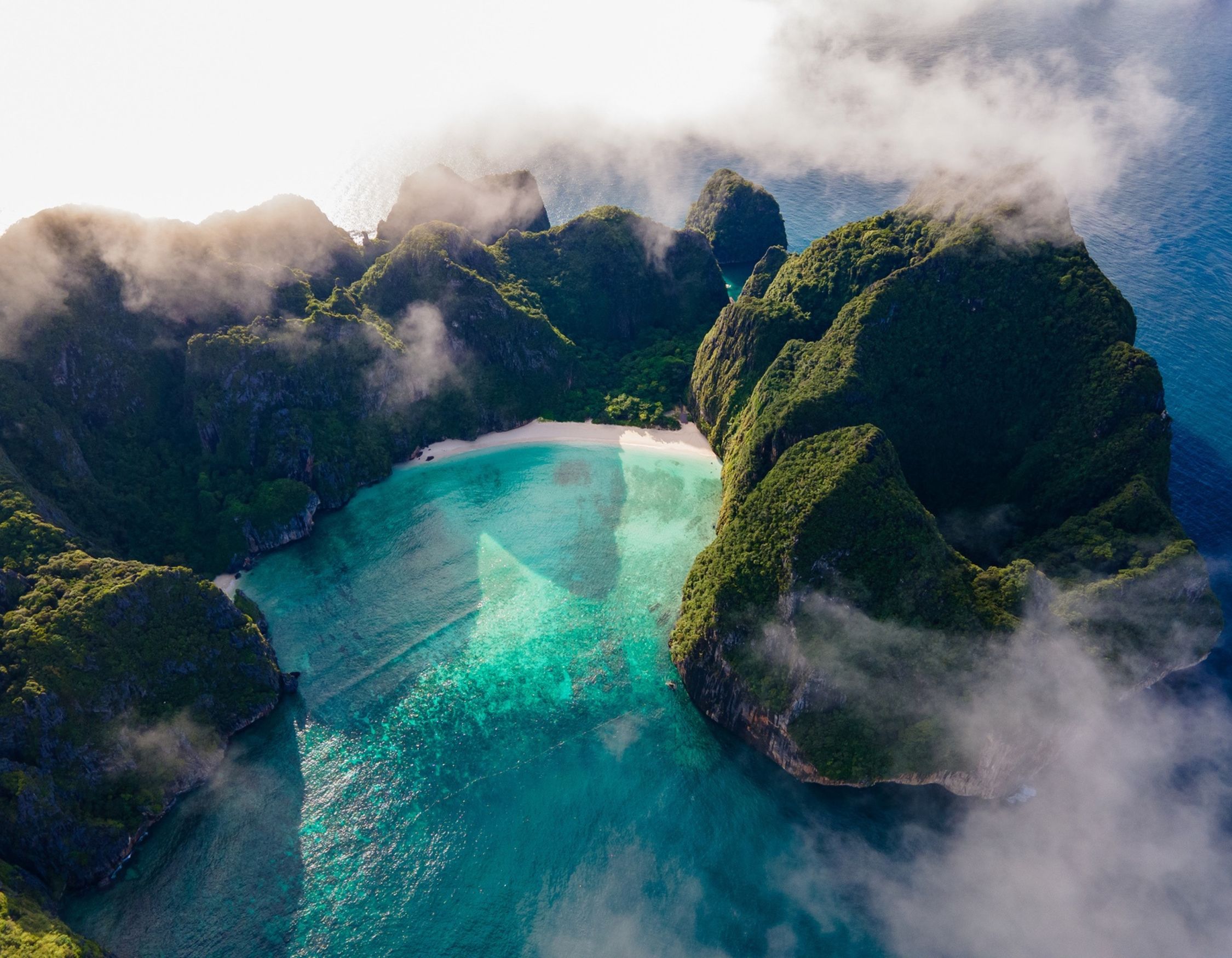 High-angle view of a tropical lagoon with turquoise water and a white sand beach enclosed by steep, misty green cliffs