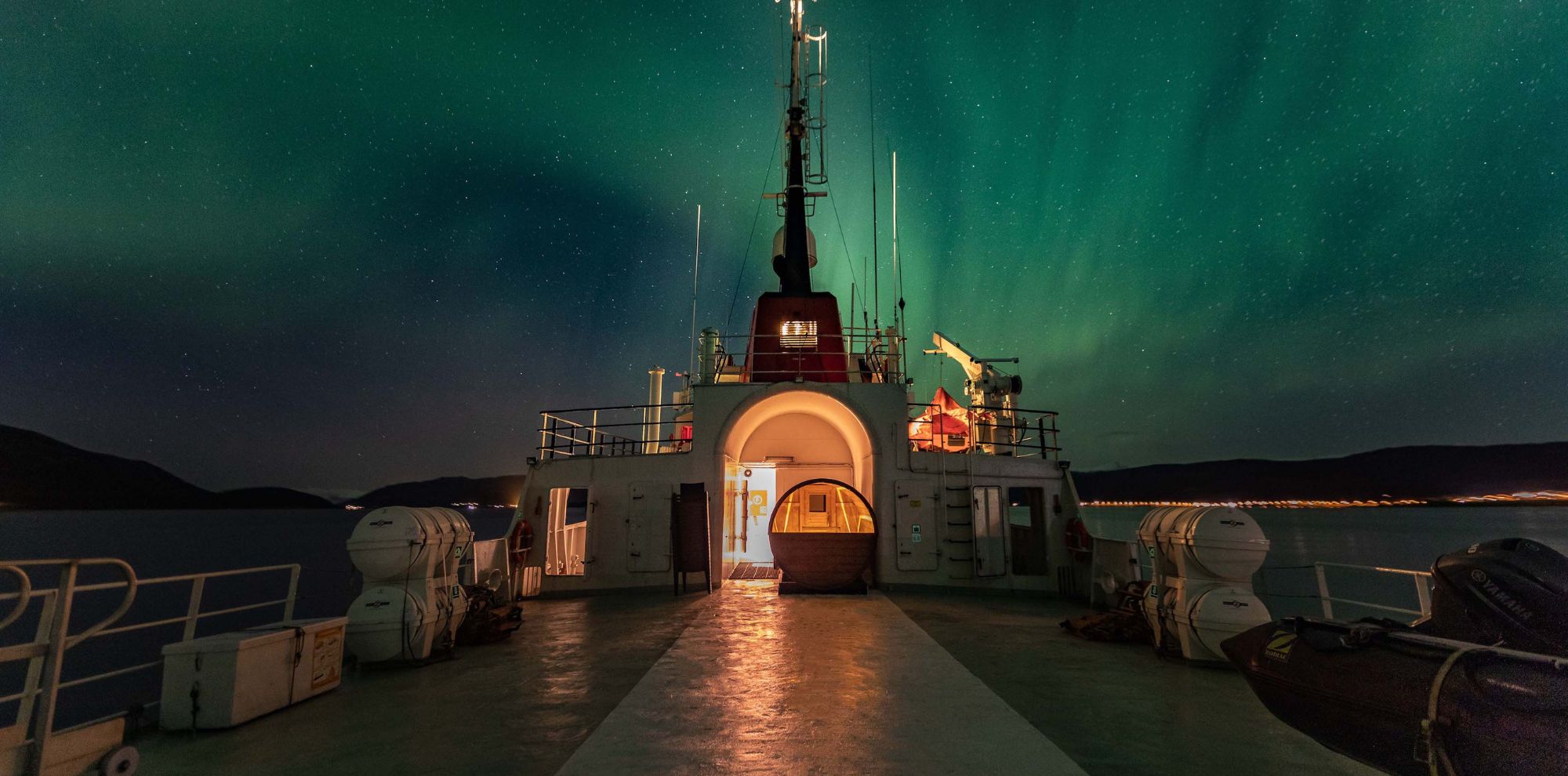 Green Aurora Borealis glows in a starry night sky above the deck of a ship with warm interior lighting.