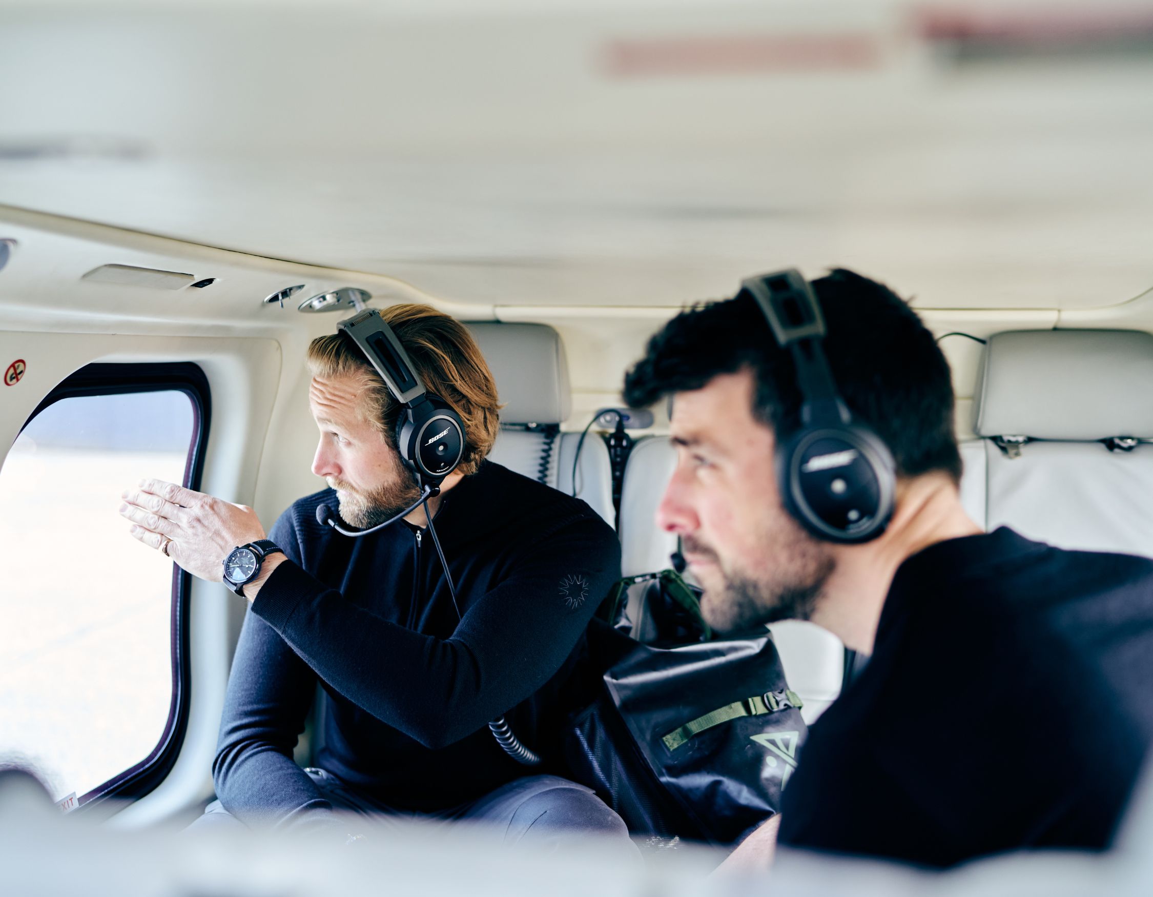 Two Pelorus Travel experts wearing headsets sit inside a helicopter, with one man pointing out of the window at the landscape below.