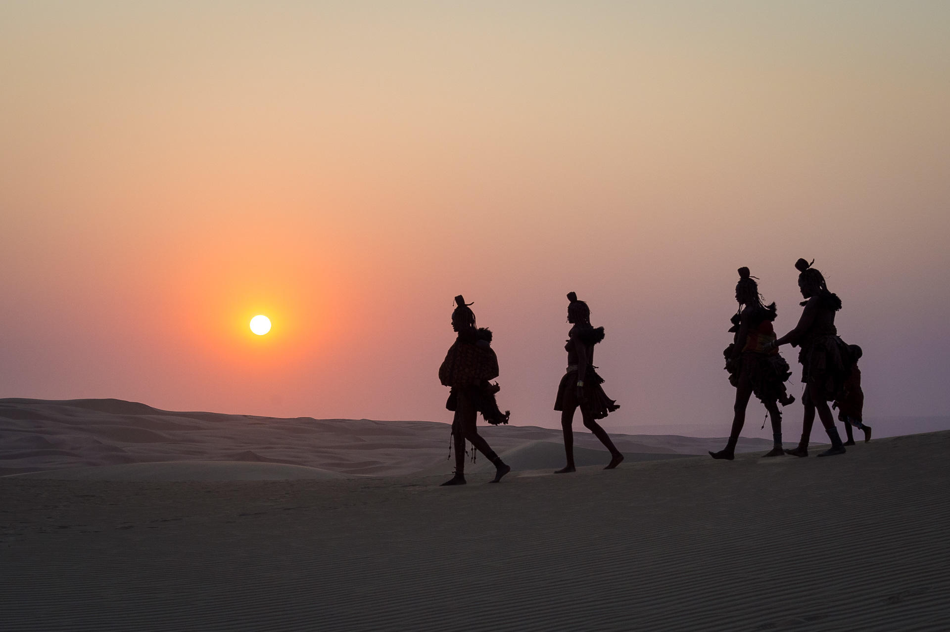 Silhouette of five people walking across a sand dune ridge toward a bright orange sun at sunset.