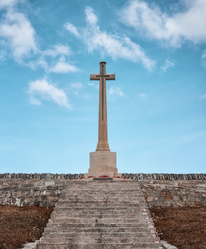 A tall stone cross stands atop a set of grey stone steps, with a blue sky and wispy clouds in the background. Small red poppy wreaths are positioned at the base of the cross.