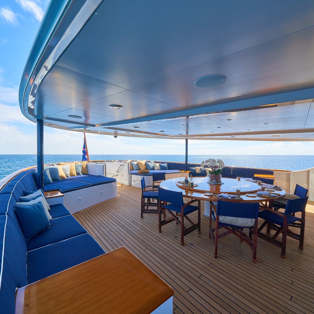 A spacious outdoor dining area on a yacht features a round table surrounded by wooden chairs. Blue cushions adorn the seating area along the edge, with a view of the ocean in the background. The deck is made of wooden planks, and the sky is clear with scattered clouds.