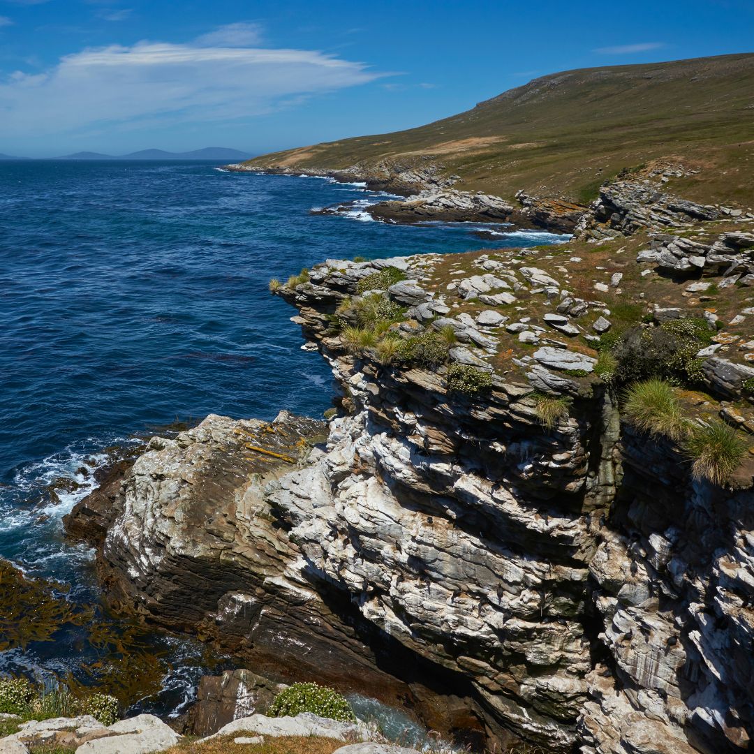 A rugged coastline with steep, rocky cliffs overlooking a deep blue sea. Lush greenery tops the cliffs, contrasting with the rocky textures. In the background, rolling hills meet the horizon under a clear blue sky.