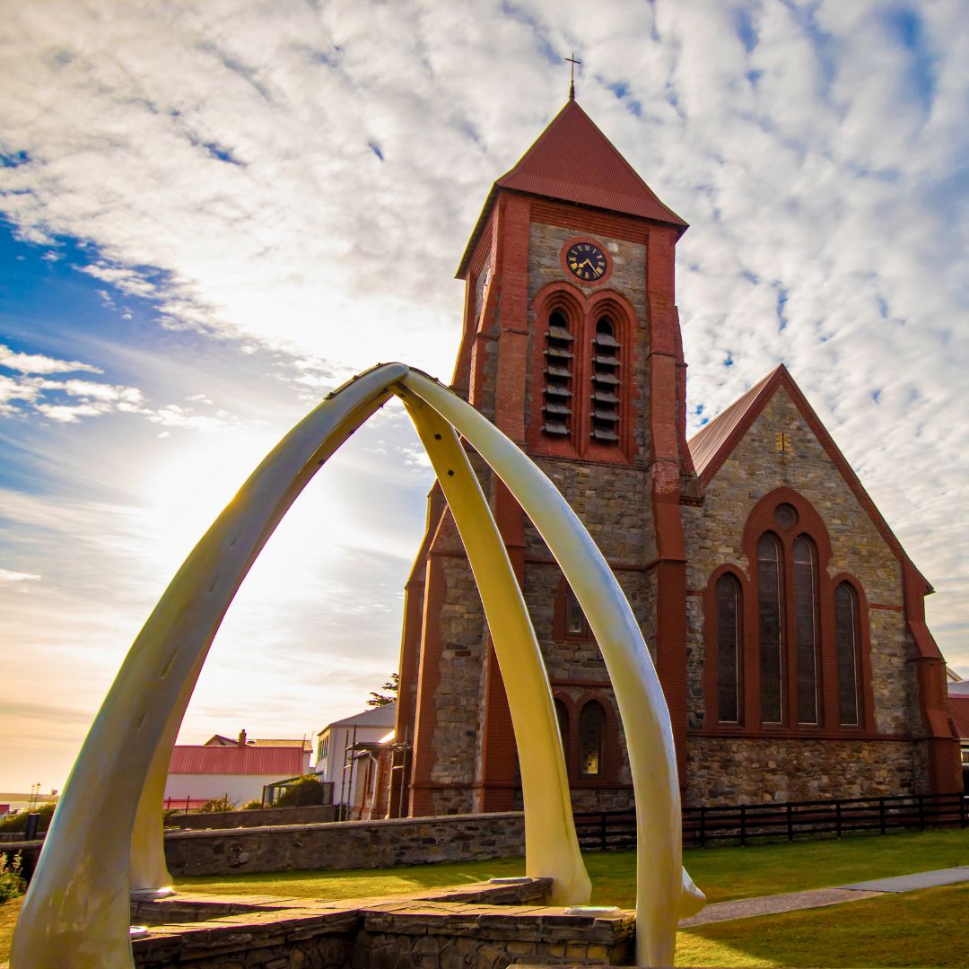A large whale bone arch frames a beautiful brick church with a clock tower, set against a backdrop of a cloudy sky and sunlight.