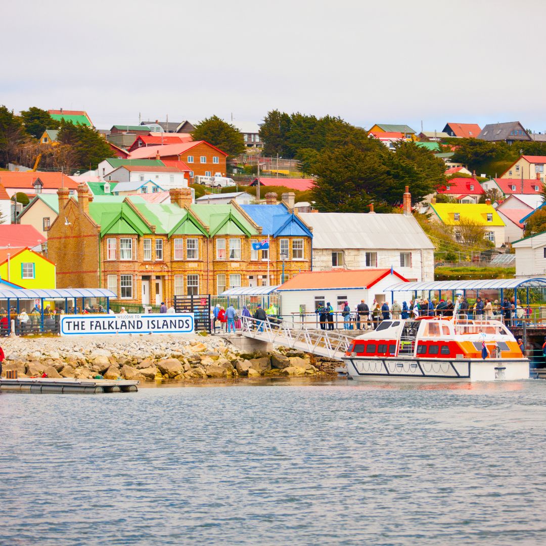 Colorful buildings line the waterfront in the Falkland Islands, with bright roofs and a bustling pier. A small boat is docked, and people are gathered along the shore, enjoying the scenic view.