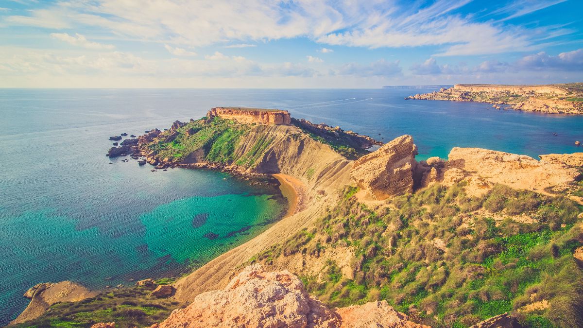 Elevated view of a rugged, grassy peninsula extending into a bright turquoise bay with rocky cliffs under a blue sky.