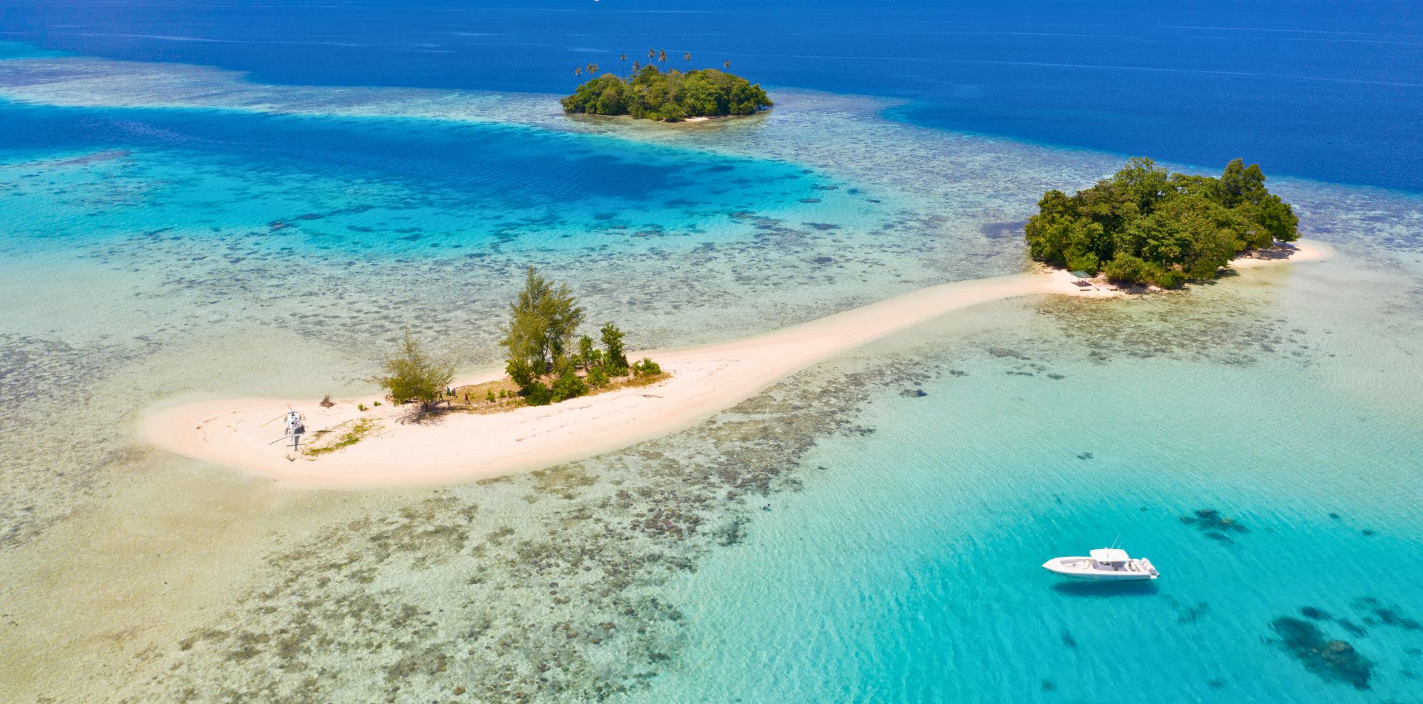 Aerial view of a tropical island with white sandy beaches and lush greenery, surrounded by clear blue waters. A small boat is anchored near the shoreline.