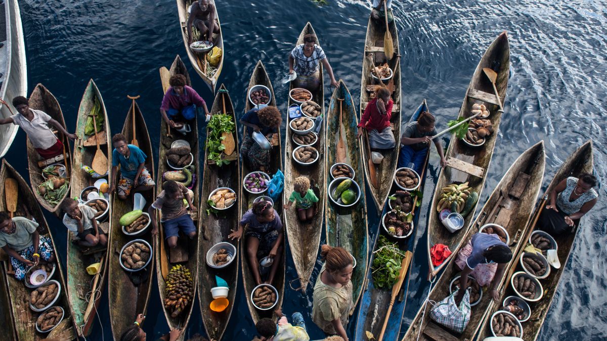Aerial view of multiple wooden boats filled with fresh produce, including fruits and vegetables, with people engaging in trade along a calm waterway.